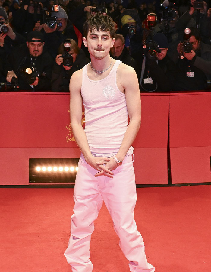 Man in white tank top and pants at Valentine’s Day red carpet, surrounded by photographers.